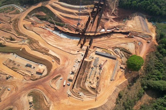 A large-scale construction site set in a partially cleared forest area, featuring winding dirt roads and an array of heavy machinery. The landscape is dominated by red-brown earth, with a linear structure running horizontally across the scene and several clusters of construction material.