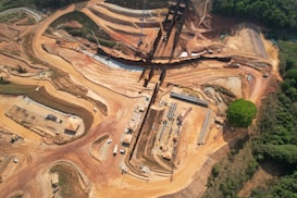 A large-scale construction site set in a partially cleared forest area, featuring winding dirt roads and an array of heavy machinery. The landscape is dominated by red-brown earth, with a linear structure running horizontally across the scene and several clusters of construction material.