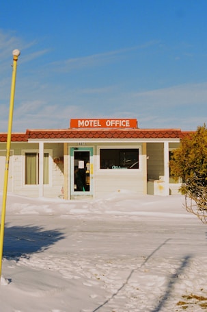 A small motel office with a brown tiled roof and a bright orange sign that reads 'Motel Office.' The building is surrounded by snow, and there is a clear blue sky. A glass door with a 'Welcome' sign is visible, and there are tracks in the snow leading to the entrance.