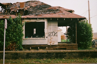 A dilapidated house with a partially collapsed roof and broken windows. The wooden siding is weathered, and graffiti is present on the front wall. Overgrown bushes surround the house, and a brick wall lines the front yard.