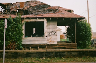 A dilapidated house with a partially collapsed roof and broken windows. The wooden siding is weathered, and graffiti is present on the front wall. Overgrown bushes surround the house, and a brick wall lines the front yard.