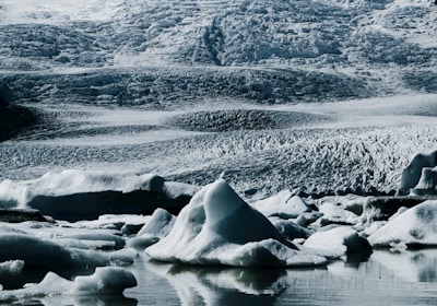 A pristine icy landscape featuring rugged glaciers and icebergs floating in calm waters. The texture of the ice suggests a cold and remote environment, with intricate patterns and layers visible on the glacier surfaces.