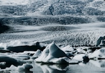 A pristine icy landscape featuring rugged glaciers and icebergs floating in calm waters. The texture of the ice suggests a cold and remote environment, with intricate patterns and layers visible on the glacier surfaces.