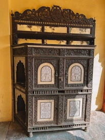 An intricately carved wooden cabinet with ornamental designs and inlaid mirror panels. The cabinet is placed against a yellow wall and has a rich antique appearance. It features detailed floral patterns and arch-shaped designs on its door panels. The top section has a decorative crest and a series of small reflective surfaces below it, showing signs of aging.