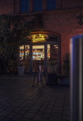 An illuminated entrance to a bar or cafe with a neon sign reading 'Food and Drinks'. The building features a brick facade with ivy growing on parts of it. The entrance is glass with visible shelves of bottles and plants inside. There is a sandwich board sign on the cobblestone path leading to the entrance. The setting appears to be in the evening, with ambient lighting providing a cozy atmosphere.