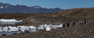 A couple dressed in wedding attire stands on a rocky shoreline with scattered icebergs floating in a calm body of water. Several other people, possibly photographers, are positioned around them capturing the scene. In the background, rugged mountains with patches of snow rise against a clear blue sky.