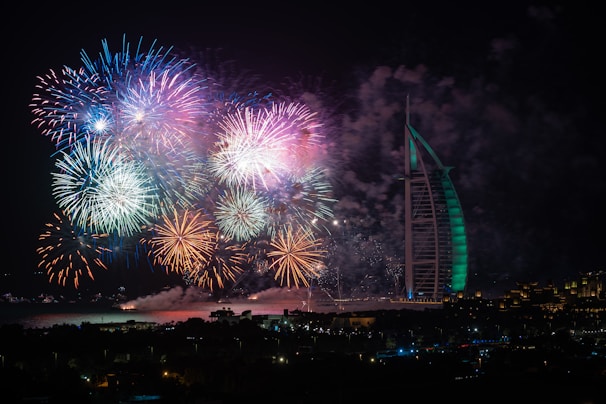 A vibrant display of colorful fireworks illuminates the night sky next to a uniquely shaped, tall building. The fireworks consist of reds, blues, greens, and whites, creating a festive atmosphere over a waterfront area with silhouetted buildings.