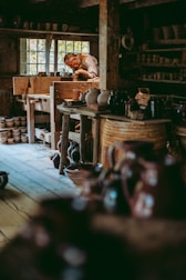 A craftsman is deeply focused on shaping pottery inside a rustic workshop filled with wooden shelves and various ceramic pieces. Natural light filters through a window, illuminating the rich textures of the wooden interior and the earthen tones of the pottery.