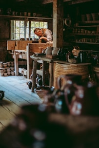 A craftsman is deeply focused on shaping pottery inside a rustic workshop filled with wooden shelves and various ceramic pieces. Natural light filters through a window, illuminating the rich textures of the wooden interior and the earthen tones of the pottery.