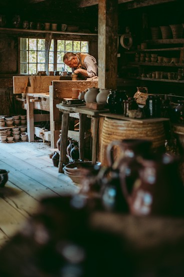 A craftsman is deeply focused on shaping pottery inside a rustic workshop filled with wooden shelves and various ceramic pieces. Natural light filters through a window, illuminating the rich textures of the wooden interior and the earthen tones of the pottery.