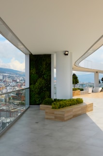 A spacious modern terrace offering expansive views of a cityscape. It features a sleek design with glass railings, potted greenery, and contemporary seating arrangements. The light-colored flooring and walls enhance the open and airy feel of the area.
