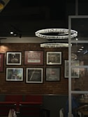 A cozy interior with a brick wall adorned with framed photographs and inspirational quotes. Two modern circular chandeliers hang from the ceiling, illuminating the space. Below, red cushioned chairs are neatly arranged against the wall. A glass partition separates this area from another section where a person is partially visible, sitting and engaged in an activity.