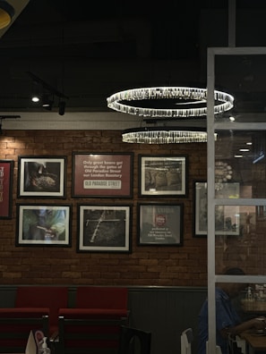 A cozy interior with a brick wall adorned with framed photographs and inspirational quotes. Two modern circular chandeliers hang from the ceiling, illuminating the space. Below, red cushioned chairs are neatly arranged against the wall. A glass partition separates this area from another section where a person is partially visible, sitting and engaged in an activity.