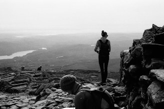 A black and white photograph captures a scenic landscape with a person standing on a rocky path, facing an expansive view of mountains and a lake in the distance. Another person is seated in the foreground, wearing a beanie, possibly engaged in a resting moment. The atmosphere appears calm and serene.
