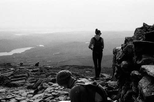 A black and white photograph captures a scenic landscape with a person standing on a rocky path, facing an expansive view of mountains and a lake in the distance. Another person is seated in the foreground, wearing a beanie, possibly engaged in a resting moment. The atmosphere appears calm and serene.