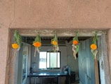 Five garlands made of marigold flowers, green leaves, and other smaller flowers hang above a doorway. The background shows an indoor area with a window, countertop, and some household items.