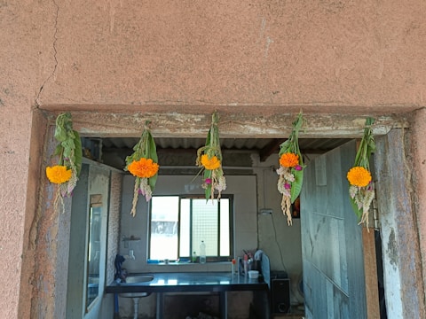 Five garlands made of marigold flowers, green leaves, and other smaller flowers hang above a doorway. The background shows an indoor area with a window, countertop, and some household items.