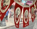 Red and white banners hanging in a row, featuring a portrait of a man with two crossed flags adorned with crescent symbols, likely representing Turkey's national flag. The word 'ATA' appears at the bottom of each banner. A blurred street scene is visible in the background, with hints of pedestrians and buildings.