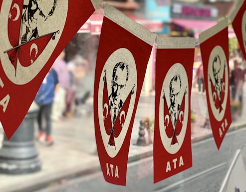 Red and white banners hanging in a row, featuring a portrait of a man with two crossed flags adorned with crescent symbols, likely representing Turkey's national flag. The word 'ATA' appears at the bottom of each banner. A blurred street scene is visible in the background, with hints of pedestrians and buildings.