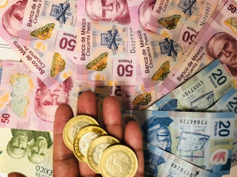 A collection of Mexican banknotes and coins is spread out. The banknotes vary in denominations and feature portraits of notable historical figures along with vibrant designs. A hand holds several gold-colored coins in the foreground.