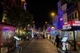 A lively street scene at night with various illuminated storefronts, restaurants, and bars on either side. The cobblestone pathway is lined with signs and menus, while several people walk down the street. Bright neon and LED lights in shades of red, blue, and yellow add vibrancy.