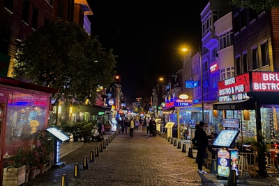A lively street scene at night with various illuminated storefronts, restaurants, and bars on either side. The cobblestone pathway is lined with signs and menus, while several people walk down the street. Bright neon and LED lights in shades of red, blue, and yellow add vibrancy.