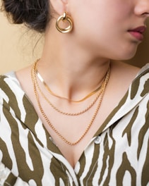 A close-up of a person's neck displaying three layers of gold necklaces and a large gold earring. The person is wearing a blouse with an animal print design in white and olive green.