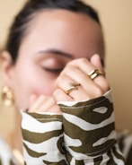 A person with closed eyes wearing gold jewelry, including a ring shaped like leaves and a pair of hoop earrings. Their hands are partially covering their face, and they are wearing a camouflage-patterned shirt.