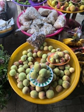 A display of fresh fruits and seeds at a market. Peaches fill a large yellow basket, with more placed on small plates. Nearby, colorful baskets hold packets of seeds or nuts. Vibrant fruits like bananas, apples, and other produce are arranged in the background.