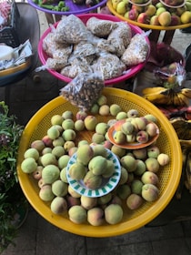 A display of fresh fruits and seeds at a market. Peaches fill a large yellow basket, with more placed on small plates. Nearby, colorful baskets hold packets of seeds or nuts. Vibrant fruits like bananas, apples, and other produce are arranged in the background.