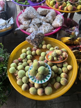 A display of fresh fruits and seeds at a market. Peaches fill a large yellow basket, with more placed on small plates. Nearby, colorful baskets hold packets of seeds or nuts. Vibrant fruits like bananas, apples, and other produce are arranged in the background.