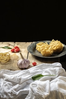 On a rustic wooden table, there are fresh pasta nests both on a metallic plate and directly on the table, along with cloves of garlic, cherry tomatoes, and green leaves, possibly basil. A white cloth is draped casually across part of the table.