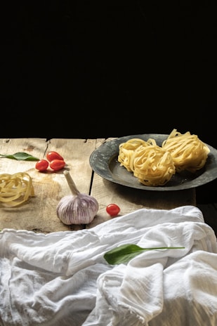 On a rustic wooden table, there are fresh pasta nests both on a metallic plate and directly on the table, along with cloves of garlic, cherry tomatoes, and green leaves, possibly basil. A white cloth is draped casually across part of the table.