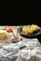 On a rustic wooden table, there are fresh pasta nests both on a metallic plate and directly on the table, along with cloves of garlic, cherry tomatoes, and green leaves, possibly basil. A white cloth is draped casually across part of the table.