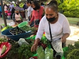 A bustling outdoor market scene featuring several women wearing face masks. A woman in the foreground is sorting or selling various fresh vegetables, including radishes and green leafy plants, which are displayed in baskets and bags. A wheelbarrow filled with dark green avocados is nearby. Other individuals can be seen interacting and purchasing goods in the background.