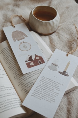 A cozy scene featuring an open book with two bookmarks placed on its pages. One bookmark displays illustrations of a sweater, an embroidery hoop, and a house, while the other has a teacup and saucer next to a lit candle with text printed on it. A small, rustic ceramic cup filled with tea or coffee sits nearby on a soft, textured fabric surface.