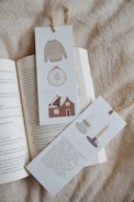 Two bookmarks with illustrations and text are placed on an open book. One bookmark features a knitted sweater, an embroidery hoop, and a house. The other showcases a cup of coffee and a candle in a holder. Both have rustic textures and are tied at the top with twine.