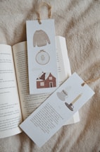 Two bookmarks with illustrations and text are placed on an open book. One bookmark features a knitted sweater, an embroidery hoop, and a house. The other showcases a cup of coffee and a candle in a holder. Both have rustic textures and are tied at the top with twine.