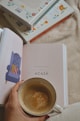 A hand holding a coffee cup with frothy coffee rests on an open book displaying an illustrated armchair and the word 'ACASĂ'. In the background, three closed books with colorful floral and bird designs are stacked.