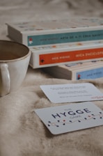 A cozy scene features a ceramic mug, a stack of books with colorful spines, and two cards on a soft, beige surface. The books have decorative covers with titles visible on the spines, and the cards are part of a game, with one card prominently displaying 'The Hygge Game.'