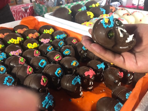 A collection of chocolate skulls adorned with colorful icing decorations, including various colors like yellow, pink, and blue. One skull is being held in a person's hand, showcasing detailed icing shapes that resemble flowers and other designs. The background contains more skulls placed on orange fabric.