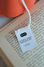 A tea tag labeled 'P&T Hunky Dory Breakfast No. 721' is placed on the pages of an open book. The text on the book page is printed in a serif typeface, and the paper appears aged. An orange object, possibly a bowl or a decorative item, is visible in the background.