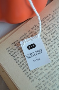 A tea tag labeled 'P&T Hunky Dory Breakfast No. 721' is placed on the pages of an open book. The text on the book page is printed in a serif typeface, and the paper appears aged. An orange object, possibly a bowl or a decorative item, is visible in the background.