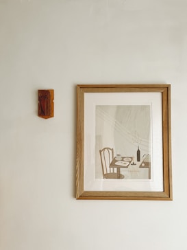 A framed artwork hangs on a plain wall, depicting a minimalistic dining scene with a table, chairs, a bottle, and tableware. To the left, a small, wooden decorative piece is mounted on the wall.