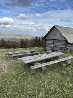 A rustic wooden cabin stands on a grassy hill with two picnic tables and benches made of logs nearby. The background features a panoramic view of rolling hills and dense forests under a partially cloudy blue sky.