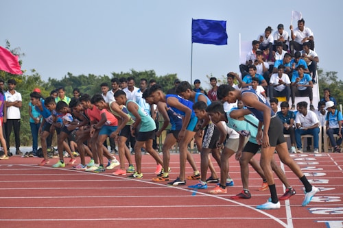A group of athletes in running attire are poised at the starting line of a track, ready to begin a race. Spectators and officials can be seen in the background, some seated on elevated platforms and others standing. The setting appears to be an outdoor athletics event.