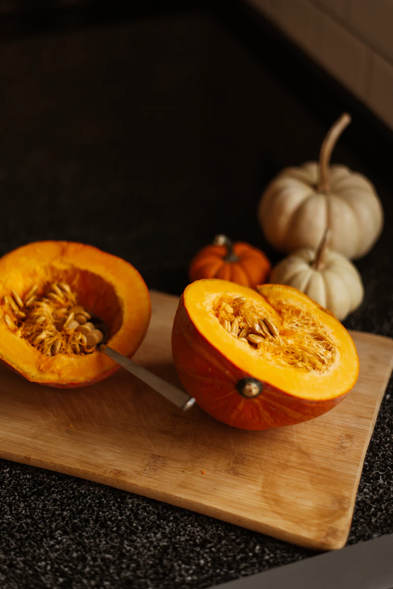 A pumpkin cut in half on a wooden cutting board, with a spoon scooping out the stringy pulp and seeds
