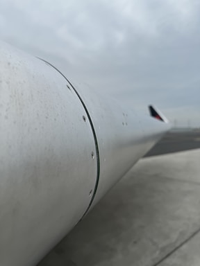 The image captures a close-up view of an airplane wing with visible rivets and a metallic finish. The background is a cloudy sky with a hint of a runway, suggesting an airport setting.