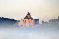 A medieval-style stone castle with tall towers and battlements is partially shrouded in mist. The castle is situated on a hilltop surrounded by light vegetation and dense fog, with hints of red and blue banners hanging down the walls. The sky above is a soft gradient, transitioning from a pale yellow to a light blue.