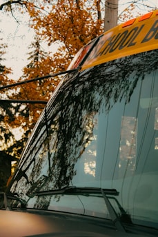 A close-up view of the front of a school bus, featuring the large windshield reflecting the surrounding autumn trees. The yellow banner above displays part of the word 'SCHOOL' in black letters. The background shows vibrant fall foliage with shades of orange and yellow.
